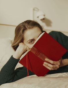 a woman laying on top of a bed holding a red book over her face with both hands