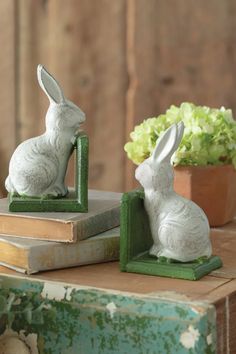 two white rabbit figurines sitting on top of books next to a potted plant