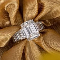 an emerald cut diamond ring sitting on top of a gold satin material baguette