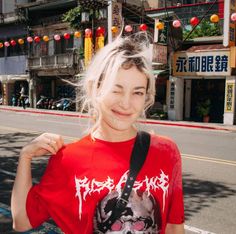 a woman standing on the side of a road with her hand in her pocket and wearing a red shirt