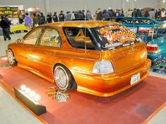 an orange car on display in a showroom with people looking at cars and other vehicles