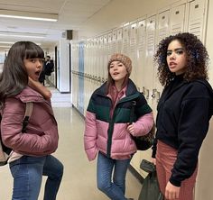 three girls standing in a hallway with lockers behind them and one girl wearing a pink jacket