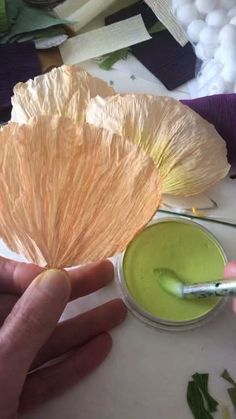 a hand holding a paper flower next to a green liquid in a small bowl on top of a table