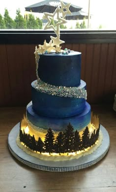 a three tiered cake with blue frosting and pine trees on the side, in front of a window