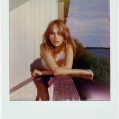 a woman leaning on a wooden bench in front of a white building with water behind her