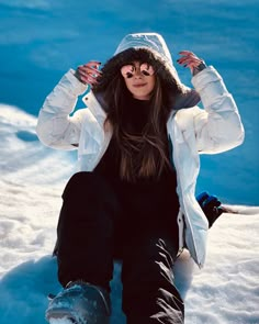 a woman sitting in the snow with her hands behind her head, wearing a white jacket and black pants