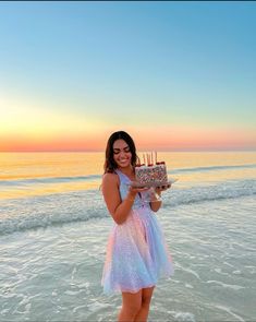 a woman standing in the ocean holding a cake with candles on it and smiling at the camera