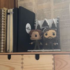 two figurines sitting on top of a book shelf next to an open book