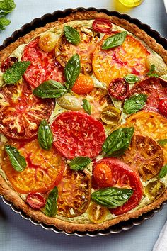 a pie with tomatoes and basil on top is sitting on a table next to lemons