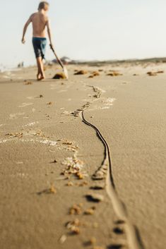 a young boy is walking on the beach with a stick in his hand and footprints in the sand