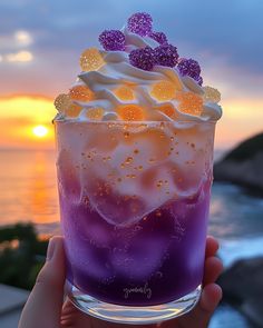 someone holding up a purple and white drink in front of the ocean at sunset or sunrise