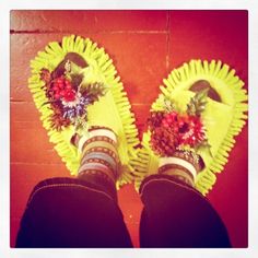 someone's feet in yellow shoes with flowers and leaves on them, standing next to a red brick wall