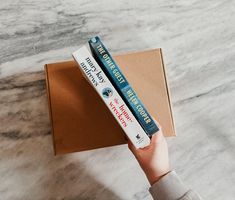 a person holding two books in front of a box on a marble counter top,