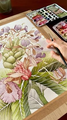 a person painting flowers on a table with paintbrushes and watercolors in front of them