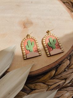 two cactus shaped earrings sitting on top of a piece of wood next to a plant