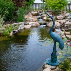 a bird statue is in the middle of a pond with rocks and plants around it