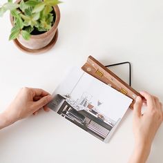 a person holding an open book on top of a table next to a potted plant