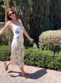 a woman in a dress is holding a water bottle and walking down the sidewalk near bushes