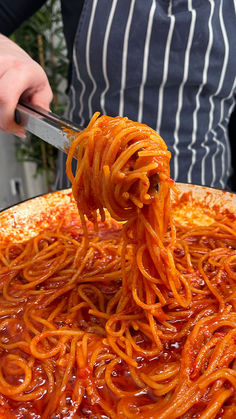 a person holding a fork full of spaghetti in front of a large pizza pie with sauce on it