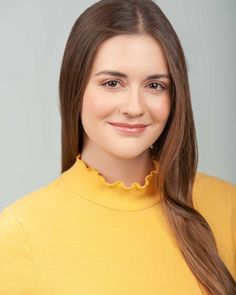 a woman with long brown hair wearing a yellow sweater and smiling at the camera while standing in front of a gray background