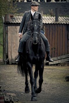 a man riding on the back of a brown horse down a dirt road next to a building