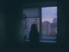 a woman standing in front of a window looking out at the city skyline from her apartment