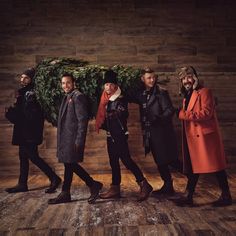 four men standing in front of a wooden wall wearing coats and hats, one man is holding a christmas tree