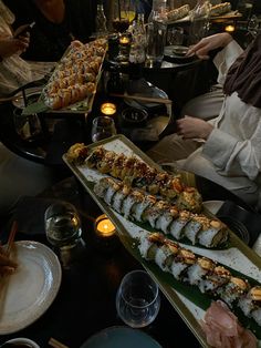 people are sitting at a table with sushi and other food items on the trays