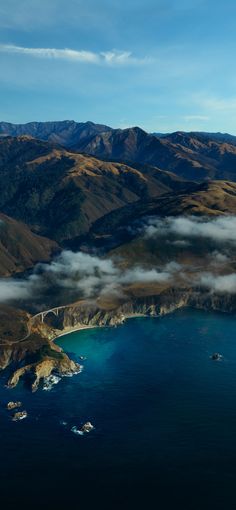 an aerial view of the ocean and mountains with clouds in the air above it is a bridge