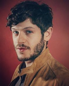 a man with curly hair and beard standing in front of a red wall wearing a leather jacket