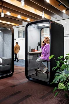 a woman sitting on a chair in an office cubicle talking on the phone while another man walks by