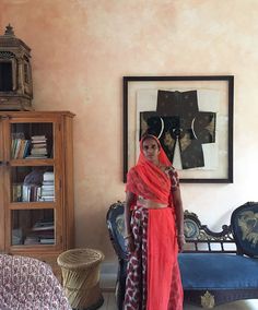 a woman in a red sari standing next to a chair and bookcase with pictures on the wall