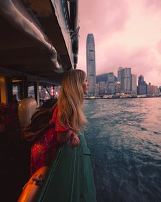 a woman standing on the side of a boat looking out at the water with buildings in the background