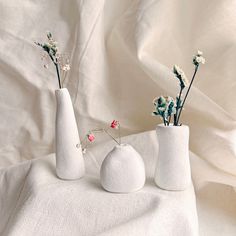 three white vases with flowers in them on a cloth covered tableclothed surface