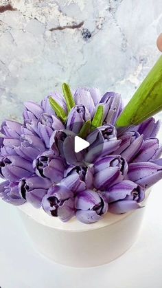 purple flowers are in a white vase on a table with a person holding the flower