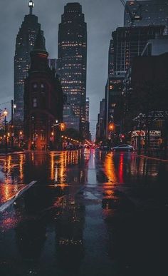 a city street at night in the rain with skyscrapers and lights reflecting off wet pavement