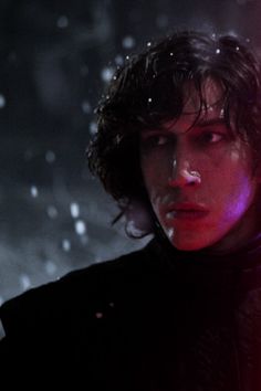 a young man with dark hair wearing a black coat and red lights behind his head