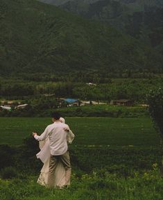 a man and woman walking through a lush green field with mountains in the back ground