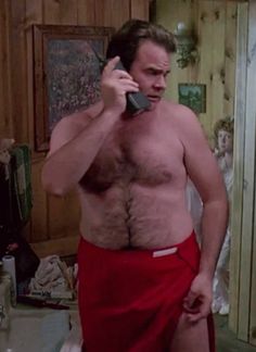 a man with no shirt talking on a cell phone in the bathroom while wearing a red towel