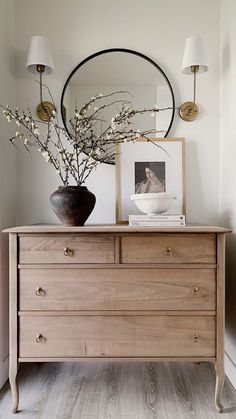 a wooden dresser topped with a mirror and vase filled with flowers next to a lamp