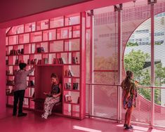 two people standing in front of a pink book shelf with books on it and another person looking out the window