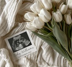 a bouquet of white tulips sitting on top of a bed next to a baby announcement