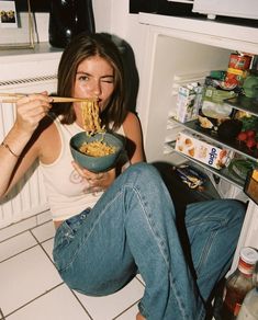 a woman sitting on the floor eating from a bowl with chopsticks in her mouth