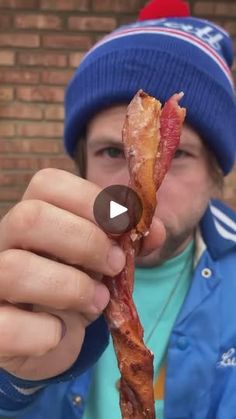 a man holding up a piece of bacon in front of his face while wearing a beanie