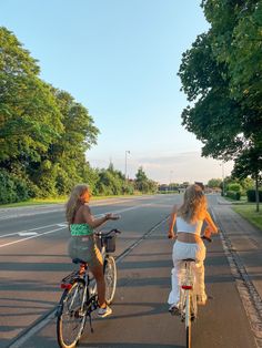 two women riding bikes on the side of a road with trees in the back ground