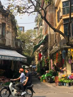 two people on a motor bike in front of an outdoor market with flowers and plants