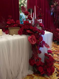 a table with red flowers and candles on it in front of a red drapes