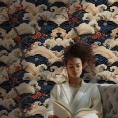 a woman sitting on a couch reading a book in front of a wallpapered background