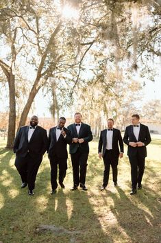 a group of men in tuxedos walking down a grass covered field next to trees