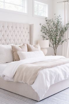 a bed with white linens and pillows in a bedroom next to a plant on the floor